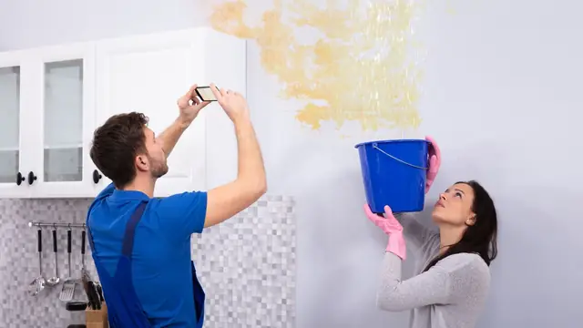 Homeowner photographing a water-stained wall while a leak is caught in a bucket